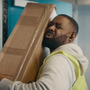 Courier man carrying large box up stairs with a pained expression on his face.